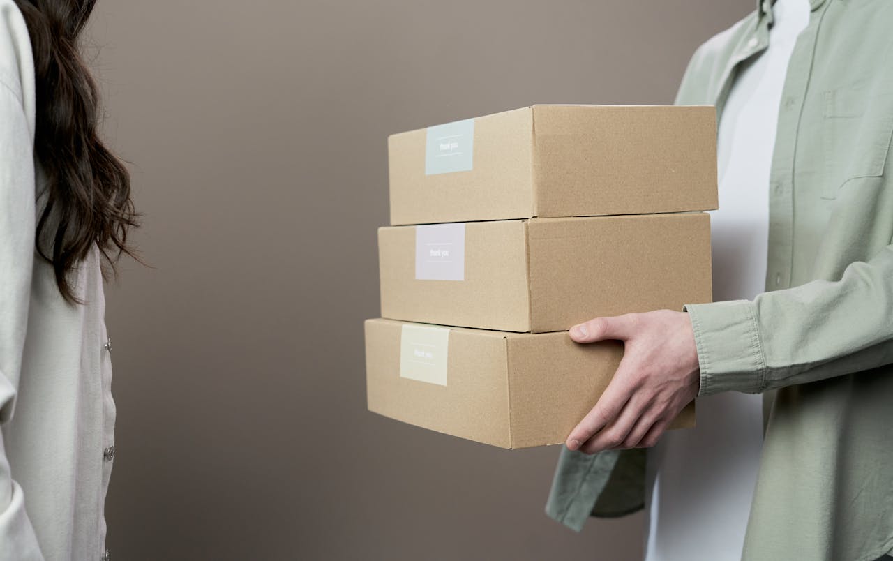 Two people exchanging stacked cardboard boxes against a neutral gray background in a studio setting.