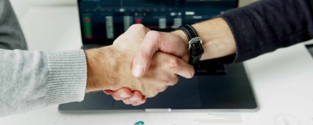 Close-up of a handshake with financial graphs on laptop screen, symbolizing a successful agreement.